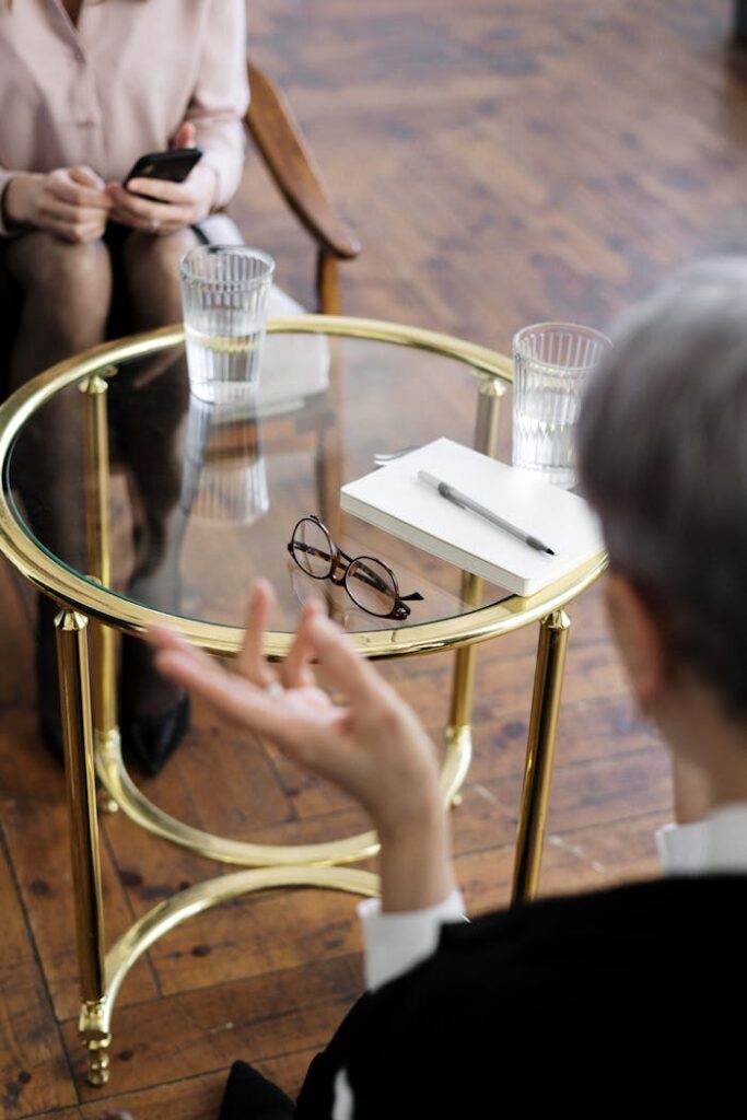 pexels-photo-4098374 Two adults engaged in a therapy session, seated at a glass table with notebooks and water glasses.